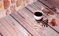 A cup of black coffee, waffle rolls, cinnamon, anise and roasted coffee beans on a wooden table against a stone wall