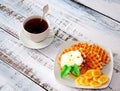 A cup of black tea and a plate with fresh waffles with cream and banana on a light wooden table