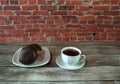 A cup of black tea on a saucer and a plate with two donuts in chocolate icing lie on a wooden table against a brick wall. Close-up Royalty Free Stock Photo