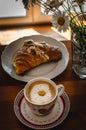 Cup of cappuccino coffee and croissant / brioche on a wooden table with freshly gathered daisy flowers. Italian breakfast. Royalty Free Stock Photo