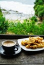 A cup of cappucino coffee in a black cup with some fried bananas in a white plate with dofocused beach dan green plants background