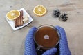 Cup of cocoa, cinnamon sticks, sugar, orange and pine cones. Hands in blue mittens holding a cup of cocoa