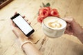 Cup of coffee in beautiful woman hands. Lady using mobile phone internet in cafe. Blank screen for layout. Red roses flowers behin