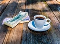 A cup of coffee and a bundle of money on a wooden table.