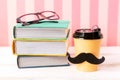 Cup of coffee with a felt moustache on the background of a stack of books and glasses. Concept of Father's day