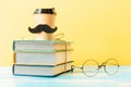 Cup of coffee with a felt moustache stands on a stack of books on yellow background with glasses. Concept of Father