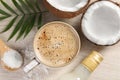 Cup of coffee, halves of coconut, flakes, bottle of syrup and green leaves on white wooden table, flat lay