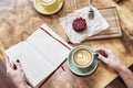 Cup of coffee latte and sweet pastry on wooden table in woman hands. hands hold opened note book, spread space for layout.