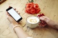 Cup of coffee and mobile phone in beautiful woman hands. Blank screen for layout. Red roses flowers behind on wooden table. St. Va