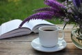 Cup of coffee, open book and bunch of purple summer flowers in glass vase on wooden table in the garden. Royalty Free Stock Photo