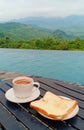 A cup of coffee and a piece of bread with a view of the poolside and mountains