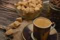 A Cup of coffee, pieces of brown sugar in a sugar bowl, coffee beans in a glass jar on a wooden background