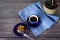Cup of coffee and pieces of homemade cake on small blue saucer on dark wooden background, napkin and flower in green ceramic pot.
