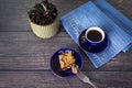 Cup of coffee and pieces of homemade cake on small blue saucer on dark wooden background, napkin and flower in green ceramic pot.