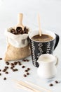 A cup of coffee with selective focus placed on the word coffee with a sack of beans in behind and cream in front.