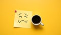 A cup of coffee sits near a note with a hand-drawn sad face on a bright yellow background Royalty Free Stock Photo