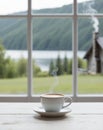 A cup of coffee on a table in front of a window with a view of a lake and log cabin. Royalty Free Stock Photo