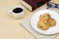 A cup of coffee, three pieces of chocolate chip cookies in a white round dish, a red book and an eyeglasses