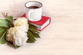 A cup of coffee, white peonies and a red book over a white wooden background.