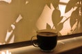 Cup of coffee on windowsill closed curtains sunrise or sunset shadows and light , crossing of plant shadows on  beige textile Royalty Free Stock Photo