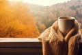 AI generated. Cup of coffee wrapped in cozy scarf and autumn leaves on windowsill with autumn park on window