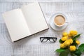 A cup of coffee, yellow peony roses and open book with glasses on the white wooden table. Flat lay. Royalty Free Stock Photo