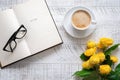 A cup of coffee, yellow peony roses and open book with glasses on the white wooden table. Time for a break. Royalty Free Stock Photo