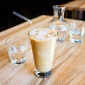 A cup of cold ice coffee with milk and tube to drink in a glass on wooden background with carafe of water Royalty Free Stock Photo