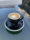 A cup of cortado coffee on a table showing the reflection of the plants in the background