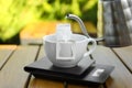 Cup with drip coffee bag, kettle and kitchen scales on wooden table, closeup