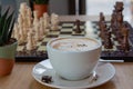 A Cup Of Frothy Coffee And Black And White Chess Pieces Lined Up On A Board