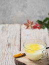 A cup with golden milk on a wooden stand. Near a wooden spoon with turmeric. Autumn leaves on a wooden background. Close-up. Copy