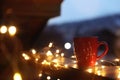 Cup of hot beverage on balcony railing decorated with Christmas lights, space for text. Winter
