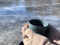 Cup of hot beverage in hands in knitted gloves across frozen lake. Outdoor activity. Winter landscape. Camping. Travel. Copy space