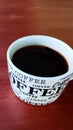 a cup of hot black coffee in a cup that says indoctrinate to love coffee always. served on a table with a bark pattern. Royalty Free Stock Photo