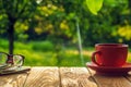 A cup of hot coffee and glasses with a newspaper on a wooden table in a green garden in the morning. Summer background Royalty Free Stock Photo