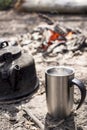 Cup with a hot drink and a kettle stand on the ground, against a blurred background of a bonfire with embers, on a sunny morning