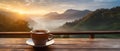 A cup of hot morning coffee with steam on a wooden table against a background of sunrise scene in the mountains.