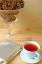 A cup of hot tea with stack of books on wooden table and a glass compote of dry pine cones in background