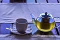 A cup of morning green tea. A transparent teapot with freshly brewed green tea and a white porcelain cup on the table by the windo