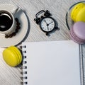 An even row of multicolored macaroons, a small alarm clock, a cup of coffee and a notebook on a wooden background. Royalty Free Stock Photo