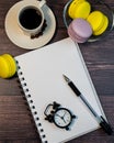 An even row of multicolored macaroons, a small alarm clock, a cup of coffee and a notebook on a wooden background. Royalty Free Stock Photo