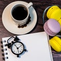An even row of multicolored macaroons, a small alarm clock, a cup of coffee and a notebook on a wooden background. Royalty Free Stock Photo