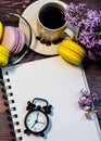 An even row of multicolored macaroons, a small alarm clock, a cup of coffee and a notebook on a wooden background. Royalty Free Stock Photo