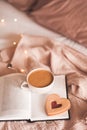 Cup of tasty coffee with red heart shape cookie on open paper book in bed over Christmas lights close up. Good morning. Breakfast Royalty Free Stock Photo