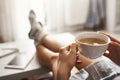 Cup of tea and chill. Woman lying on couch, holding legs on coffee table, drinking hot coffee and enjoying morning