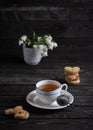 A cup of tea with cookies and a tea strainer on a dark background with bunches of white flowers of snowdrops