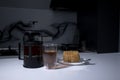 cup of tea in a double-botoom glass, french-press teapot and piece of cake on white table in gray kitchen