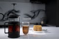 cup of tea in a double-botoom glass, french-press teapot and piece of cake on white table in gray kitchen