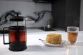 cup of tea in a double-botoom glass, french-press teapot and piece of cake on white table in gray kitchen